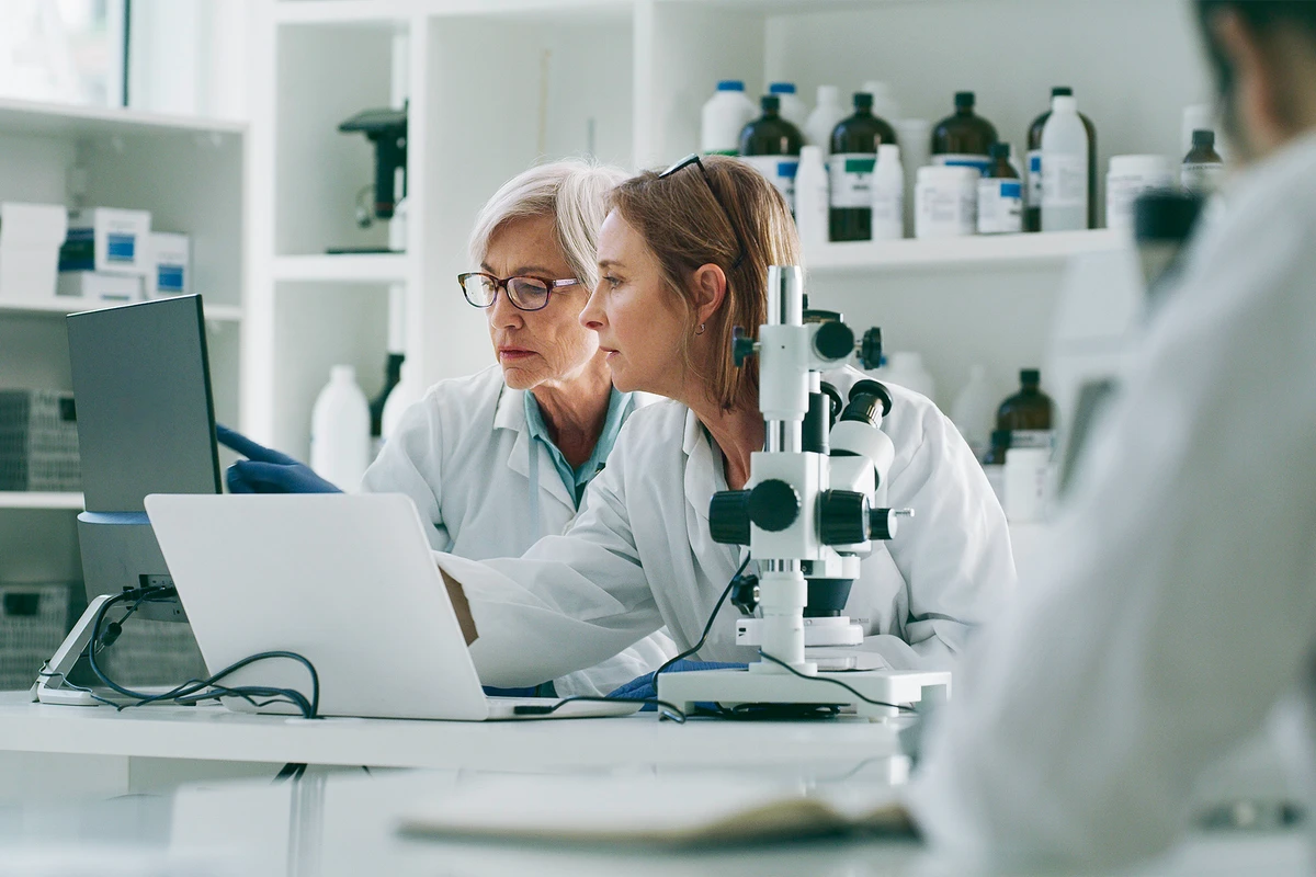 Zwei Wissenschaftlerinnen analysieren gemeinsam Labordaten an einem Computer neben einem Mikroskop.