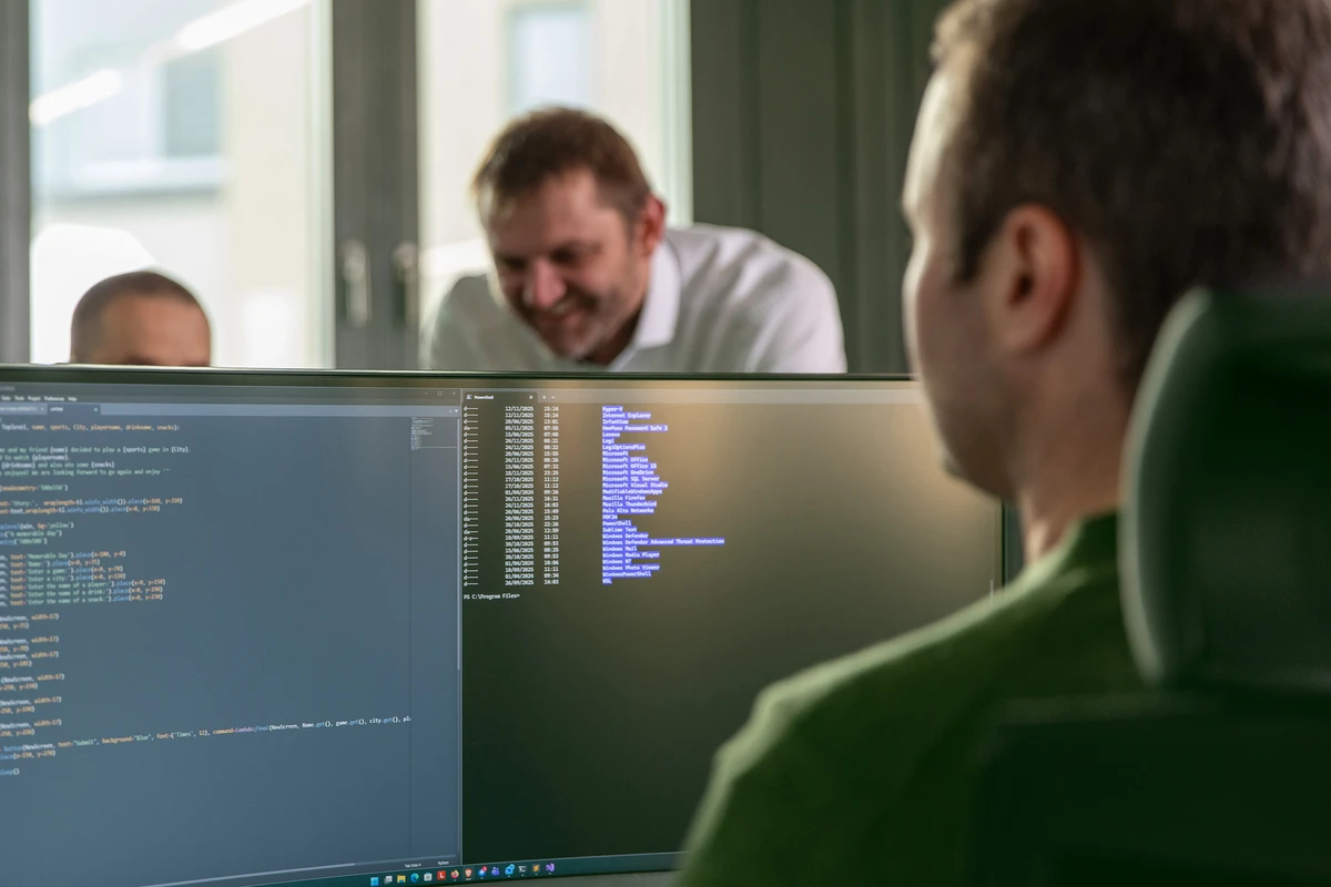 Zwei Personen besprechen sich im Hintergrund am Schreibtisch und eine Person sitzt am Schreibtisch davor.