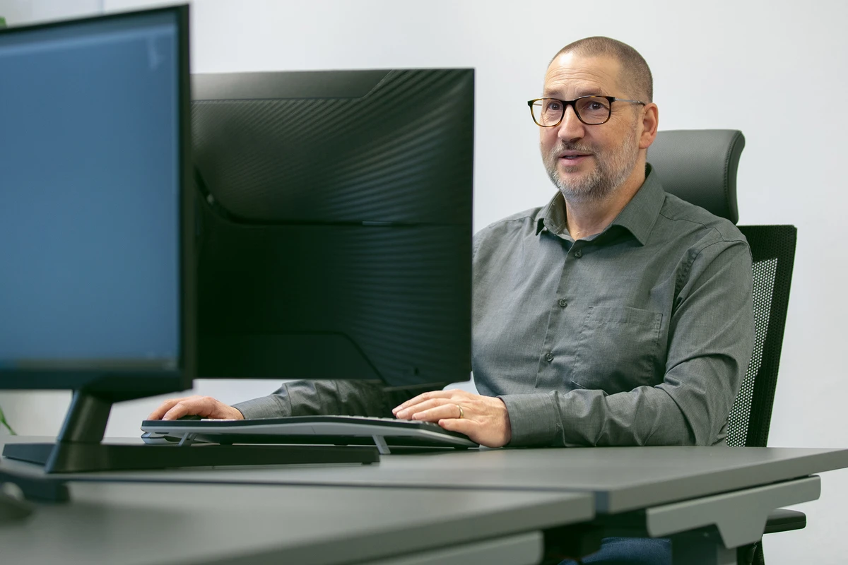 Person sitzt an einem Schreibtisch vor einem großen Monitor und arbeitet am Computer.
