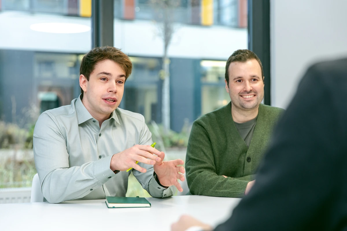 Zwei Personen sitzen nebeneinander in einem Meeting und sprechen mit der Person im Vordergrund.