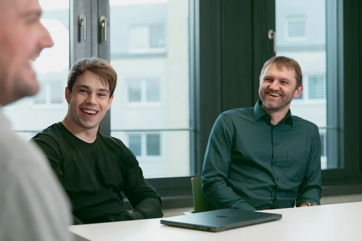 Starke Teamkultur für erfolgreiches IT- und Data Consulting. Drei Personen sitzen an einem Tisch und lächeln amüsiert während eines Meetings.