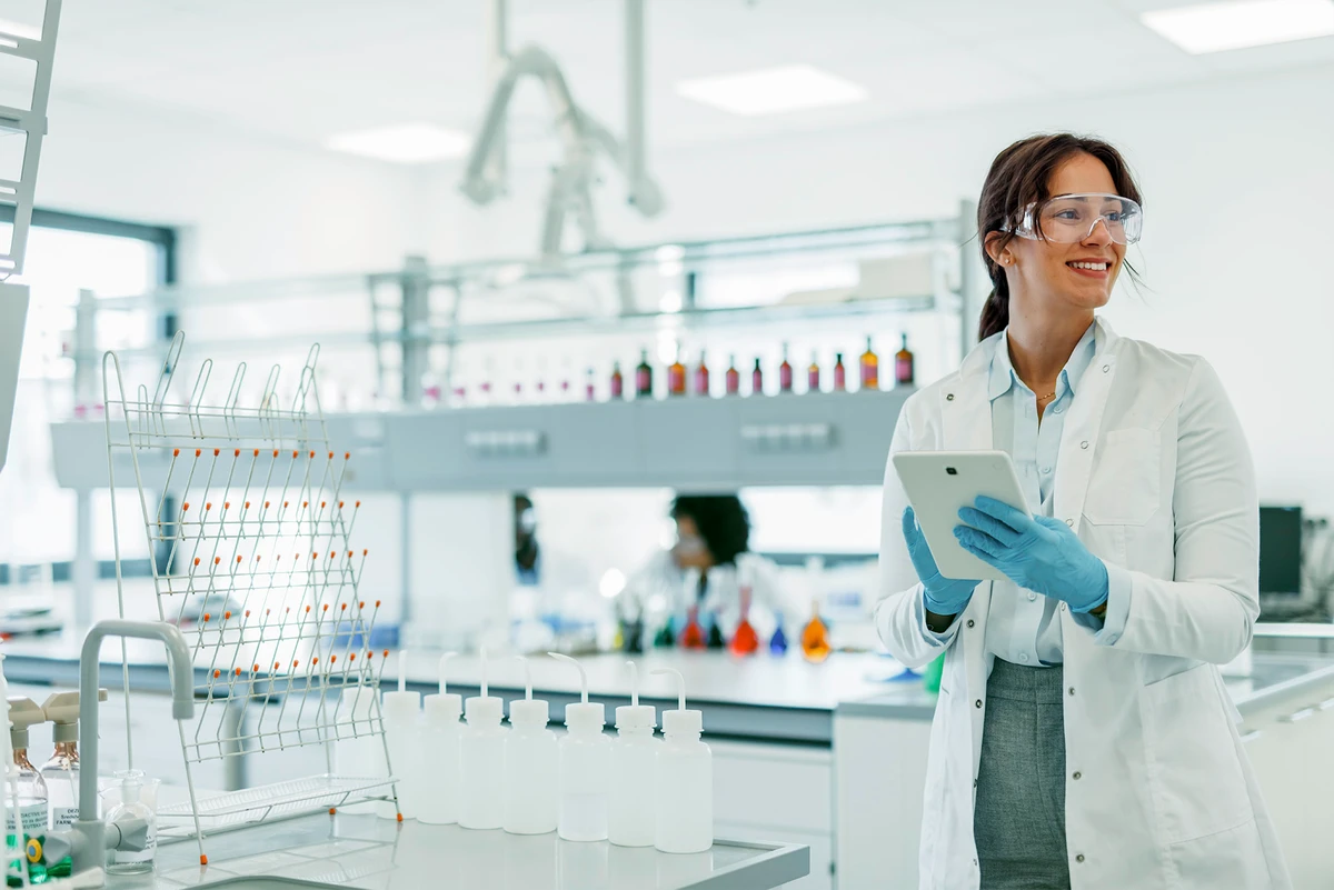 Wissenschaftlerin mit Tablet und Schutzbrille steht in einem modernen Labor zwischen Laborarbeitsplätzen.