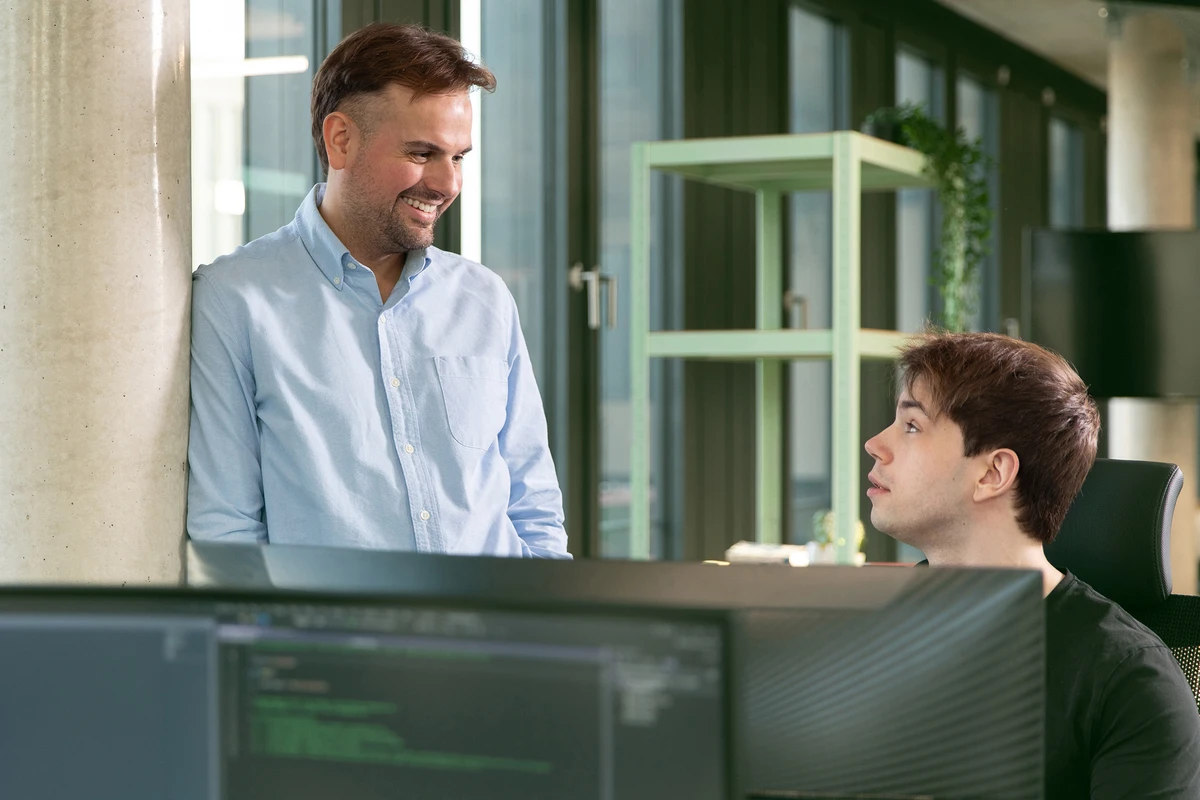 Eine Person steht und tauscht sich mit seinem Kollegen vor einem Rechner über ein Projekt aus.